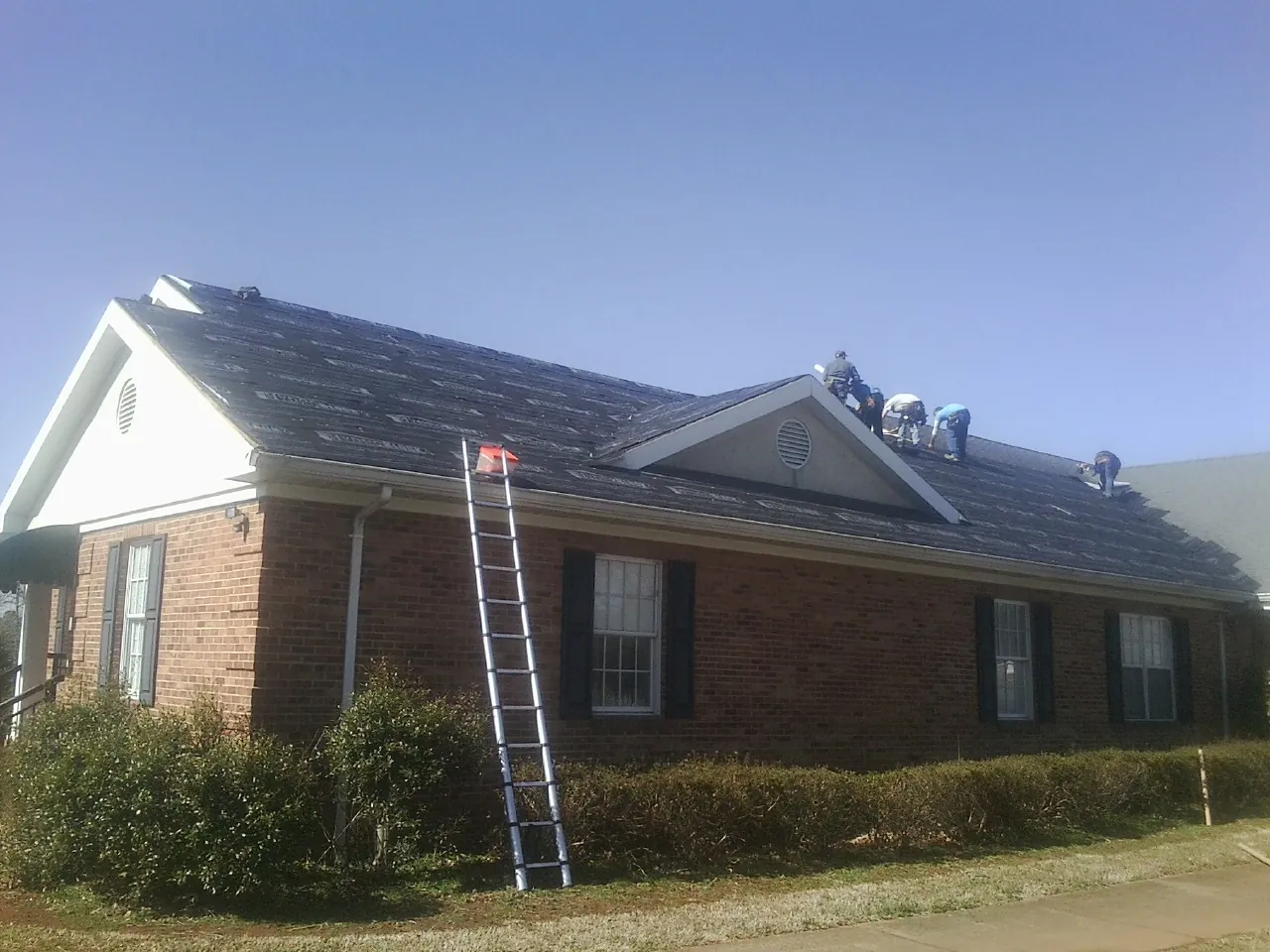 Crew tearing shingles off a brick ranch roof with a ladder set — Metal Roof Repair work by our crew in Ocean Isle Beach