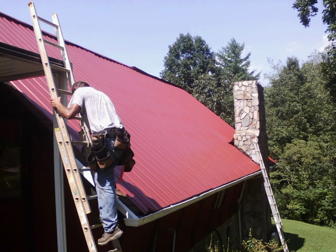 Roofer on a ladder beside a red metal roof and stone chimney — Soffit & Fascia Repair by Roofing Brothers in Ocean Isle Beach